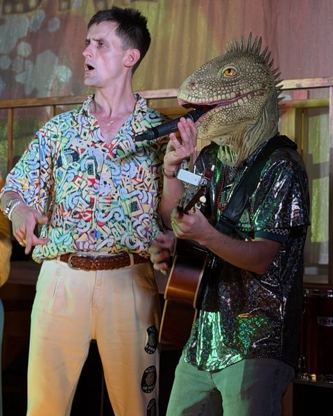 Performing live on stage: a singer in a patterned shirt beside a guitarist in a lizard mask